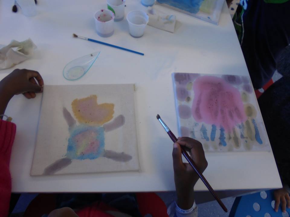 Mãos de aluno com pincel e dois quadros pintados com as tintas fabricadas