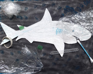 Trabalho final: o tubarão em relevo feito em cartolina sobre um fundo negro e rodeado de lixo marinho - plástico transparente, um cotonete, uma espátula em plástico, um bocado de rolha de garrafa de plástico.