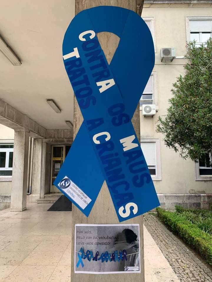 Laço azul gigante à entrada do Hospital com o texto "Contra os maus tratos a crianças" junto a uma folha A4 com lacinhos presos e o texto "Por nós, pelo fim da violência, leve-nos consigo!"