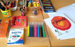 Materiais de pintura como lápis de cor e de cera e canetas de feltro dispostos em cima da mesa junto do livro "O ponto".
