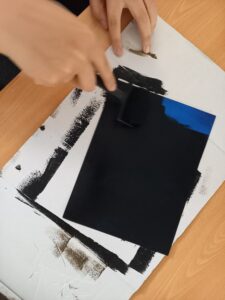 Uma aluno pinta com um rolo a cor negra sobre a tela que já tem outras cores pintadas previamente.