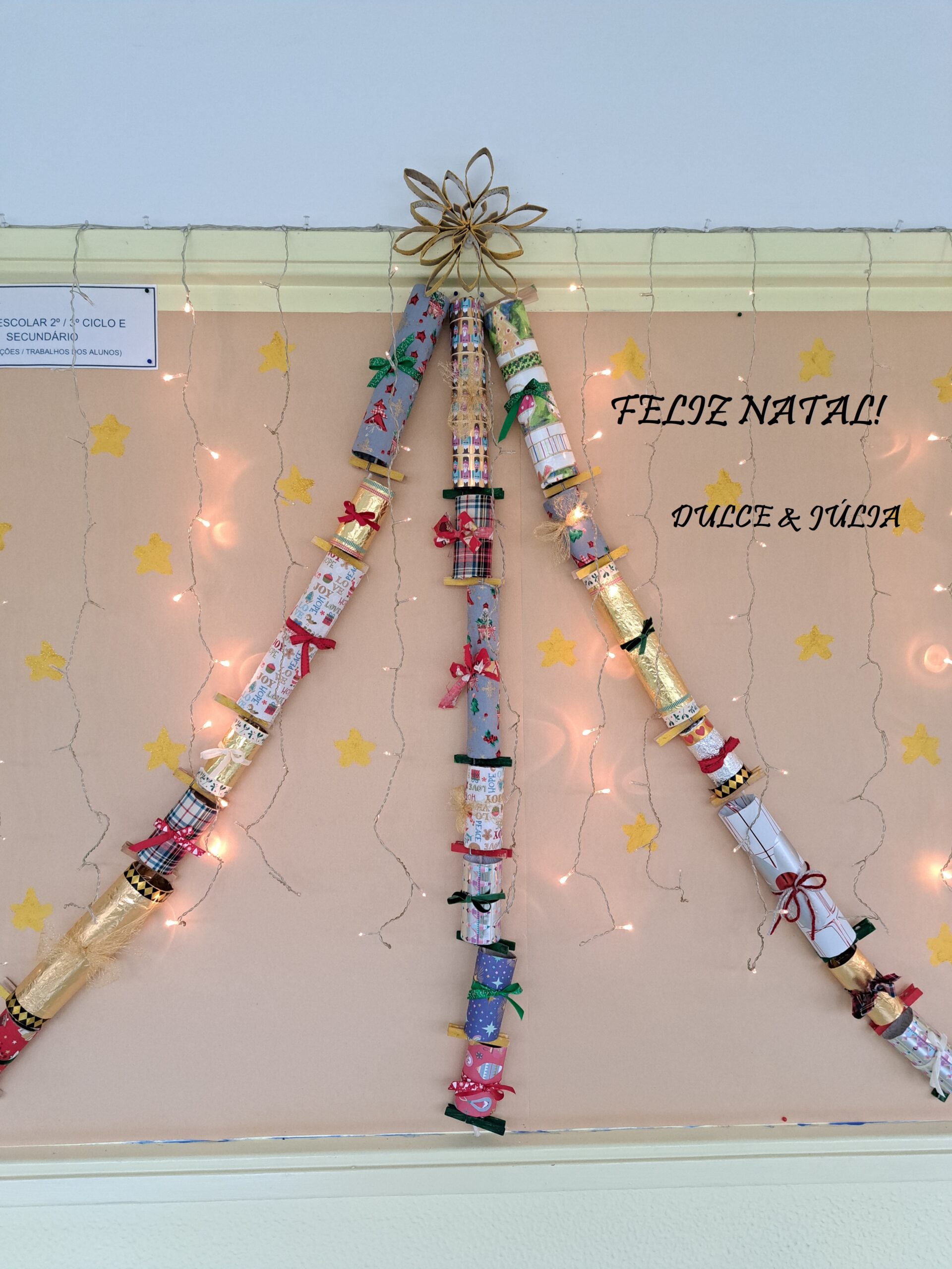 Tubos multicoloridos numa parede dispostos de modo a fromarem uma Árvore de Natal; texto "Feliz Natal! Dulce e Júlia"